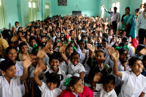 CDSP Photo taken in Kolkutta, India. Compassion's Sensitive Messaging Standards apply to this asset for external usage. See Compassion's Communications Manual (p. 22-26) https://maxplus.compassion.com/viewpicture.tlx?pictureid=22119320320 for the standards. View of a large inside, indoors, crowded classroom, class, of children, boys and girls, youth, students, classmates, looking up smiling happily and waving with their hands, while teachers, center staff, adult men and women, tutors, stand along the side painted blue walls.